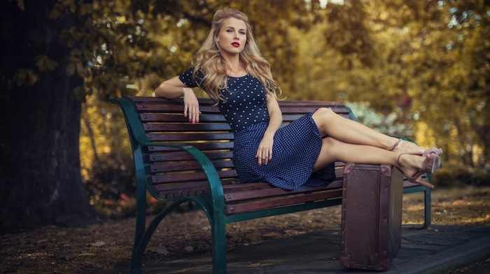 blonde, looking at viewer, bench, blue dress, model, long hair, girl outdoors, red lipstick, dress, suitcases, polka dots, depth of field, high heels, sitting, leaning, girl